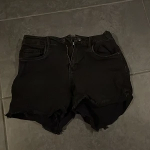 Svarta jeansshorts - Säljer ett par svarta jeansshorts i bra skick. De har en rå kant och är perfekt för sommarens alla äventyr. Shortsen har en dragkedja och knappar framtill samt fickor både fram och bak. Passar perfekt för en avslappnad stil!