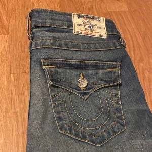 Blå jeans från True Religion - Säljer ett par snygga blå jeans från True Religion i bra skick. De har en bootcut-stil och är tillverkade i Mexico. Jeansen har coola detaljer som kontrastsömmar och en unik fickdesign med knapp. Perfekta för både vardag och fest!