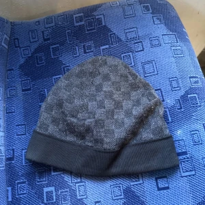 Grå mössa från Louis Vuitton - Snygg grå mössa från Louis Vuitton med ett diskret rutigt mönster. Den har en uppvikt kant och en liten LV-logga framtill. Perfekt för att hålla sig varm med stil under kyliga dagar. Passar både till vardags och lite finare tillfällen.