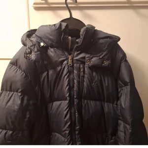 Dunjacka från Ralph Lauren - Säljer en snygg och varm mörk blå dunjacka från Ralph Lauren. Jackan har en broderad logga på bröstet och dragkedja framtill. Perfekt för kalla vinterdagar med sin pufferstil och långa ärmar. Den är i bra skick. Om ni har en annan jacka ni vill byta kan ni även skriva