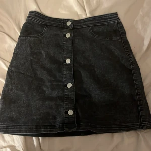 Svart jeanskjol från H&M - Snygg svart jeanskjol från H&M i storlek 14+. Kjolen har en rad med knappar framtill och en klassisk jeanslook. Perfekt för både vardag och fest! 🖤