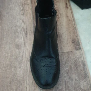 Svarta skinnstövletter med dragkedja - Säljer ett par snygga svarta stövletter i skinn. De har en stilren design med perforerade detaljer på tån och en praktisk dragkedja på sidan för enkel på- och avtagning. Perfekta för både vardag och fest!