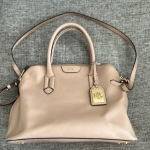Beige handväska från Ralph Lauren - Säljer en superfin beige handväska från Ralph Lauren. Den är i skinn och har både handtag och en axelrem, vilket gör den perfekt för både vardag och fest. Väskan har en dragkedja upptill och en snygg RL-tag på framsidan. Insidan är rymlig med en extra ficka för småsaker. Perfekt för dig som vill ha en stilren och praktisk väska!