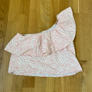 Rosa och vit zebramönstrad oneshoulder topp från MNG - Säljer en superfin rosa och vit zebramönstrad off shoulder topp från mango. Toppen har en volangdetalj som ger den en extra touch av stil. Perfekt för sommarens fester eller en dag på stan. Den är i nyskick och redo att bäras av någon som älskar att sticka ut!
