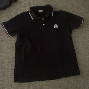 Svart pikétröja från Moncler - Säljer en snygg svart pikétröja från Moncler. Den har korta ärmar och en klassisk krage med röd och vit rand. Tröjan har två knappar framtill och Monclers logga på bröstet. Perfekt för en stilren look!