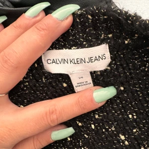 Svart stickad tröja från Calvin Klein Jeans - Säljer en snygg och bekväm svart stickad tröja från Calvin Klein Jeans. Tröjan har ett diskret mönster med små inslag av guldtrådar som ger den en unik touch. Den är långärmad och perfekt för höst och vinter. Passar både till vardags och fest. Tröjan är i mycket bra skick och knappt använd. Perfekt för att hålla sig varm och stilren under de kallare månaderna. 🖤✨