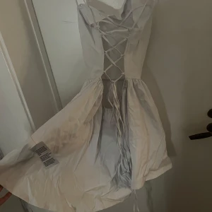 Vit studenrklänning med snörning - Säljer en söt vit klänning med smala axelband och snörning i ryggen. Klänningen har en kort längd och är perfekt för varma dagar. Den har en enkel och stilren design som gör den lätt att matcha med olika accessoarer. Lite liten i storleken 