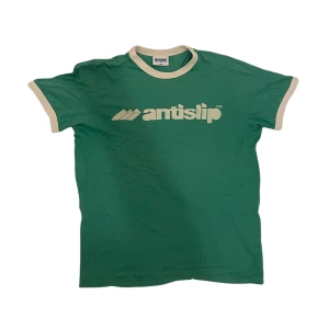 Grön Antislip t-shirt - Snygg grön t-shirt från Antislip med vitt tryck på framsidan. T-shirten har vita kanter vid ärmar och hals some ger en retro känsla. Perfekt för en avslappnad stil. Igenkligen storlek L men passar mer som S kanske lite M.
