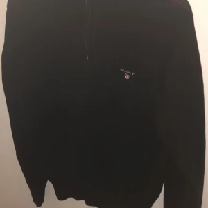 Svart tröja med dragkedja från Gant - Säljer en stilren svart tröja från Gant med dragkedja och ribbad krage. Perfekt för kyligare dagar och passar till det mesta. Tröjan har en liten logga på bröstet och är tillverkad i ett mjukt material.