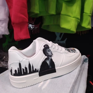 Vita sneakers med New York-motiv - Unika vita sneakers med tryck av en stadssilhuett och porträtt. Texten 'New York' pryder framsidan med en flaggdetalj. Skorna har klassisk snörning och en stilren design.
