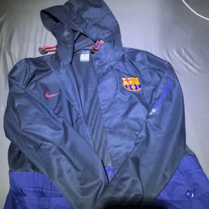 Mörkblå barcelona vindjacka/rengjacka - Säljer en mörkblå vindjacka från Nike med huva och dragkedja. Jackan har en broderad logga på bröstet och en ficka med dragkedja på sidan. Perfekt för sportiga tillfällen eller som en lätt ytterjacka.