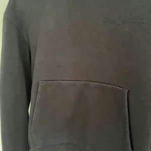 Svart hoodie från Sail Racing - Säljer en svart hoodie från Sail Racing med en stor ficka framtill. Tröjan har långa ärmar och en diskret logga på bröstet. Perfekt för en avslappnad stil.