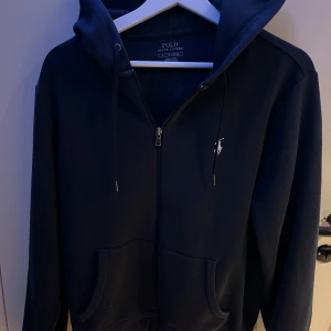 Mörkblå hoodie från Ralph Lauren - Säljer en stilren mörkblå hoodie från Ralph Lauren med dragkedja och klassisk logga på bröstet. Den har en justerbar huva med snören och praktiska fickor framtill. Perfekt för en avslappnad look. pris kan diskuteras vid snabb affär