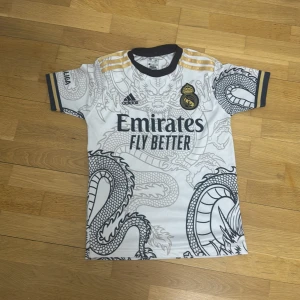 Ronaldo legend tröja real Madrid  - Snygg vit fotbollströja från Adidas med ett coolt drakmönster i grått. Tröjan har korta ärmar och guldiga detaljer på axlarna. Perfekt för fotbollsfans som vill sticka ut på planen!