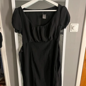 Svart klänning med knytband - Elegant svart klänning med korta puffärmar och knytband i ryggen. Tillverkad i Italien, perfekt för en stilren look. Klänningen har en rundad halsringning och en smickrande passform.