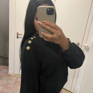 Svart tröja med guldknappar - Snygg svart tröja med dekorativa guldknappar på axeln. Tröjan har en normal passform och långa ärmar, perfekt för en stilren look. Passar bra till både jeans och kjol.