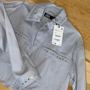 Ljusbgrå jacka från Zara - Snygg ljusgrå jacka från Zara med silverfärgade nitar på krage och fickor. Jackan har en stilren design med knappar framtill och två bröstfickor. Perfekt för att ge din outfit en cool touch. storkek XS 