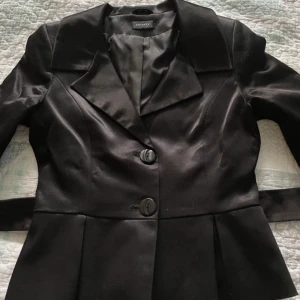 Office  siren  Svart kavaj från Onyx - Elegant svart kavaj från Onyx med knappar framtill och ett matchande bälte i midjan. Kavajen har en klassisk krage och långa ärmar, perfekt för en stilren look.