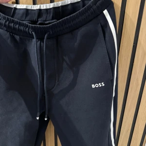 Svarta mjukisbyxor från Boss - Snygga svarta mjukisbyxor från Boss med vita detaljer längs sidorna. Byxorna har en elastisk midja med snörning och en bakficka. Perfekta för en avslappnad stil. Storlek Xs från room 1006 från emporia. Lite nopriga bak och vid inre sidan vid midjan annars jätte fint skick inga fläckar eller hål. Skulle säga färgen är mörkblå/svartz