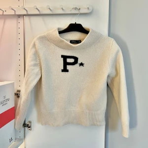 Vit/Beige stickad tröja från Polo Ralph Lauren - Säljer en stilren vit/beige stickad tröja från Polo Ralph Lauren med ett stort marinblått 'P' och stjärna på framsidan. Det är en halv polo!! Tröjan har långa ärmar och ribbade kanter vid ärmslut och nederkant. Perfekt för en avslappnad och trendig look. OBS vet att storleken säger ”L” men den har krympt i tvätten så det är en M!! Självklart äkta, köpt i Paris förra året för ca 3000. Den har inga skador utom att den är lite nopprig men jag tar bort det med noprringsmaskin innan jag skickar! 