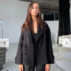 Svart kappa från Design by Si - Snygg svart kappa från Design by Si med dubbelknäppning och bred krage. Jackan har långa ärmar med dekorativa knappar vid ärmsluten. Perfekt för kyliga vår/höst dagar! 