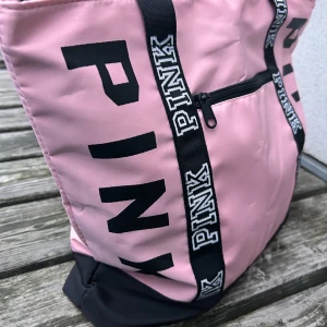 Rosa totebag från PINK! - Snygg rosa totebag med svarta detaljer i slitstark canvas. Väskan har en praktisk dragkedja framtill och rymligt inre för alla dina saker. Perfekt för en dag på stan, skolan eller som gymväska. Mått: 40 x 40 cm. Ny och oanvänd. 