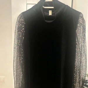 Svart sammetklänning med paljettärmar - Elegant svart klänning i sammet från Lamar Fashion. Den har långa, transparenta ärmar dekorerade med glittriga paljetter som ger en festlig touch. Perfekt för en glamorös kväll!