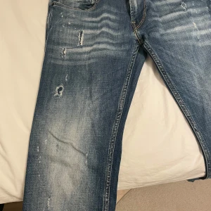 Blå jeans från Replay - Snygga blå jeans från Replay med slitna detaljer och en klassisk blå färg. Jeansen har används litegrann bara. Värt att notera att högra framfickan är sönder alltså ingen ficka men det är simpelt att laga!