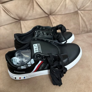 Svarta sneakers med vita sulor - Snygga svarta sneakers med vita sulor och textdetaljer på sidan. Skorna har snörning och en stilren design med röda och blå ränder. 