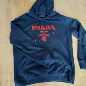 Svart hoodie från Prada - Säljer en svart hoodie från Prada med röd broderad logga på framsidan. Tröjan har en klassisk design med huva och dragsko samt en stor ficka framtill. Perfekt för en stilren och bekväm look.Äkta