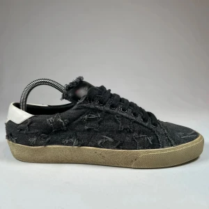 Svarta sneakers från Saint Laurent - Snygga svarta sneakers från Saint Laurent i jeansmaterial med slitna detaljer. Skorna har en vit häl och svarta snören. Perfekta för en avslappnad stil med en touch av lyx.