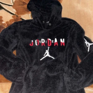 Svart Jordan hoodie - Säljer en svart Jordan hoodie med det ikoniska Jumpman-logotypen och texten 'JORDAN' i rött och vitt på framsidan. Hoodien har en mjuk och fluffig yta med dragsko i huvan. Perfekt för en sportig och avslappnad stil.