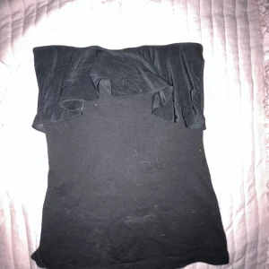 Svart tubtopp med volangdetalj - Snygg svart tubtopp med volangdetalj upptill. Perfekt för en stilren och enkel look. Passar bra till jeans eller kjol för en avslappnad stil.