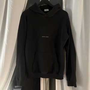 Svart hoodie från Saint Laurent - Snygg svart hoodie från Saint Laurent med diskret logga på bröstet. Tröjan har en klassisk design med huva och en stor ficka framtill. Perfekt för en stilren och avslappnad look.