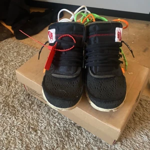 Nike x Off-White sneakers - Säljer ett par svarta Nike x Off-White sneakers med unika detaljer som röd tag och flerfärgade skosnören. Skorna har en modern design med vit sula och Off-White branding på sidan.