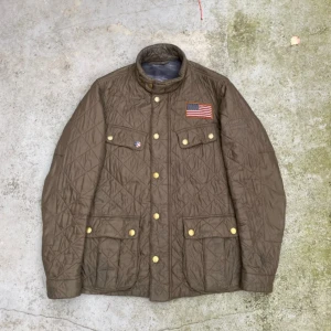 Olivgrön quiltad jacka från Barbour - Säljer en olivgrön quiltad jacka från Barbour International Steve McQueen-kollektionen. Jackan har en amerikansk flagga på bröstet och flera praktiska fickor. Perfekt för höst och vår.         Pris kan diskuteras