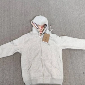 Grå huvtröja från Burberry - Säljer en stilren grå huvtröja från Burberry med dragkedja och broderad logga på bröstet. Insidan av huvan har det klassiska Burberry-rutiga mönstret. Perfekt för en avslappnad och trendig look.