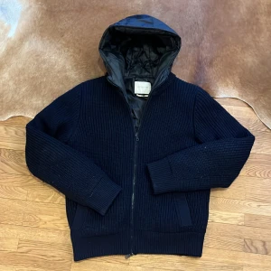 Brooksfield stickad cardigan hoodie - Marinblå stickad jacka av ullblandning. Jackan har en quiltad insida för extra värme. Liten skada som syns på sista bilden men inget som märks av, annars toppkvalite på jackan och skicket. M - L i storlek. Nypris: 429 euro ~4900 kr. Perfekt för våren! 