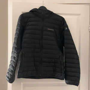 Svart dunjacka från Peak Performance - Snygg svart dunjacka från Peak Performance med huva och dragkedja. Perfekt för kyliga dagar. Jackan har en stilren design med diskret logga på bröstet och ärmen. Jackan är lite sliten under vänster armbåge men annars felfri