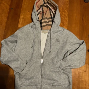 Grå Burberry hoodie - Säljer en stilren grå hoodie från Burberry med dragkedja och klassiskt rutigt foder i huvan. Tröjan har en broderad logga på bröstet och är tillverkad i bomull med en liten del polyester för extra komfort. Perfekt för en avslappnad stil.