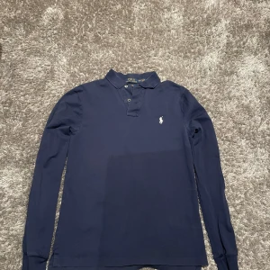 Mörkblå pikétröja från Ralph Lauren - Säljer en klassisk mörkblå pikétröja från Ralph Lauren med lång ärm och den ikoniska loggan broderad på bröstet. Perfekt för en stilren look. Tröjan har en krage och knappar framtill.