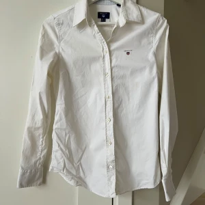 Vit skjorta från Gant - Säljer en klassisk vit skjorta från Gant med broderad logotyp på bröstet. Skjortan har långa ärmar och knappar framtill. Perfekt för en stilren look.
