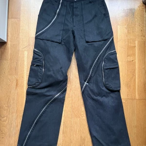 AW 2003 Dolce & Gabbana spiral jeans - Aw 2003 dolce Gabbana spiral jeans. Snygga svarta byxor från Dolce & Gabbana med unika diagonala dragkedjedetaljer och praktiska cargofickor. Perfekta för en stilren och modern look. Byxorna har en normal passform och är tillverkade i ett slitstarkt material. Sista bilden är inte min, vill bara visa vad andra säljer för.