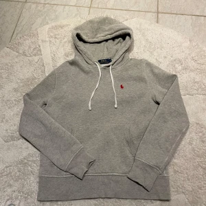 Grå hoodie från Ralph Lauren - Säljer en stilren grå hoodie från Ralph Lauren med den klassiska loggan broderad i rött på bröstet. Tröjan har en justerbar huva med vita snören och ribbade muddar vid ärmslut och nederkant. Perfekt för en avslappnad look. 14-16