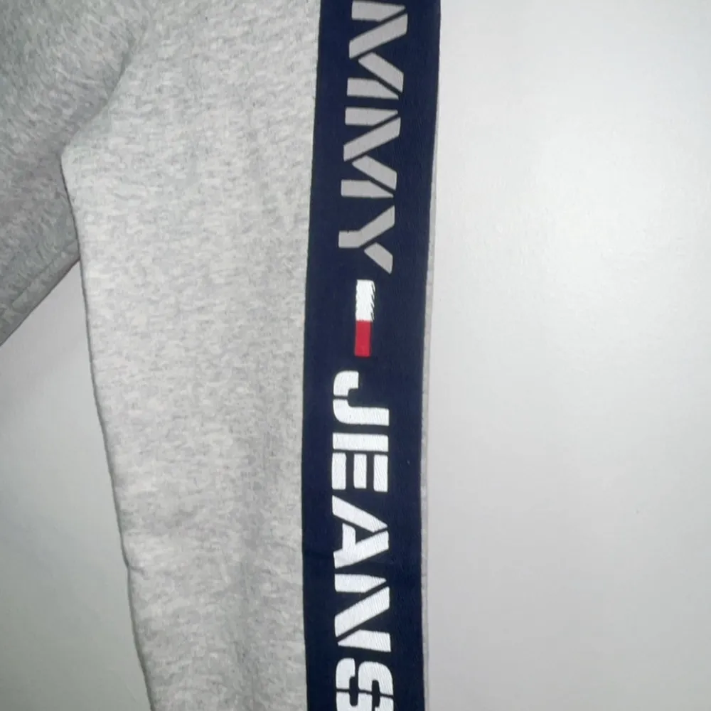 Säljer en snygg grå sweatshirt från Tommy Jeans. Den har långa ärmar med mörkblå detaljer längs sidorna och märkesloggan tryckt på ärmen. Tröjan är i bomull och har ribbade muddar vid ärmslut och nederkant. Perfekt för en casual look!. Tröjor & Koftor.