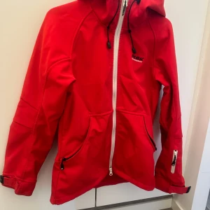 Röd jacka från Peak Performance - Säljer en snygg röd skaljacka från Peak Performance. Den har en dragkedja framtill och justerbara snören vid huvan. Vindtät, vattenavstötande. Jackan har praktiska fickor med dragkedjor och är i bra skick. 