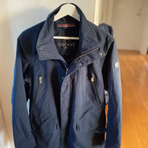 Mörkblå jacka från Snoot - Säljer en snygg mörkblå jacka från Snoot i storlek S. Jackan har en stilren design med dragkedja och knappar framtill. Perfekt för vår och höst med sin lätta och bekväma passform. Den har långa ärmar och praktiska fickor med dragkedjor. En klassisk och tidlös stil för alla tillfällen.