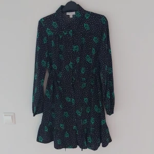Svart och grön klänning från Topshop - Säljer en superfin mörkblå klänning från Topshop med gröna blommor och vita prickar. Den har långa ärmar och en söt volangdetalj längst ner. Perfekt för både vardag och fest!