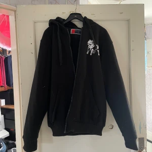 Svart hoodie med hundmotiv från V-T Fashion - Säljer en svart hoodie från V-T Fashion i storlek L. Den har ett coolt hundmotiv i vitt på både framsidan och baksidan. Hoodien är gjord i ett mjukt och mysigt material, perfekt för kyliga dagar. Den har en dragkedja framtill och en stor huva. Perfekt för en avslappnad stil!