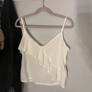 Vit ärmlös topp med volangdetalj - Säljer en superfin vit ärmlös topp med volangdetalj framtill. Perfekt för en festlig kväll. Toppen har smala axelband och en luftig passform. Den är gjord i ett lätt och bekvämt material. Volang tvärs över💕💕